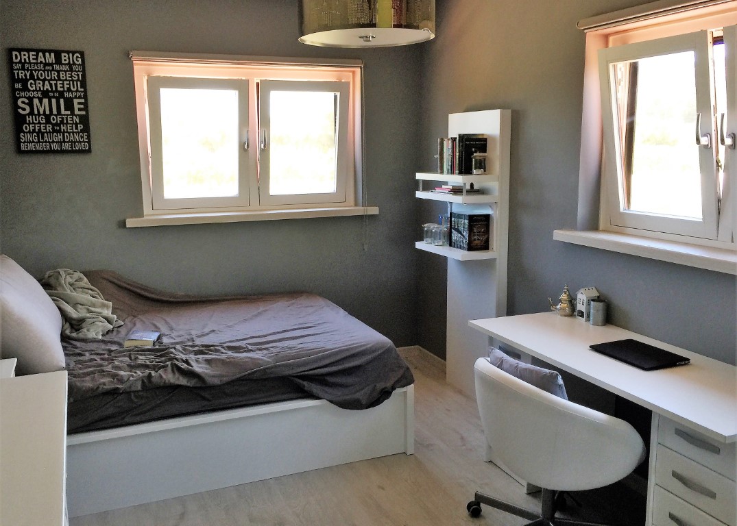 Doppelzimmer mit integriertem Arbeitsbereich, grauen Wänden und hellem Holzboden. Zwei Fenster sorgen für reichlich natürliches Licht im Raum.