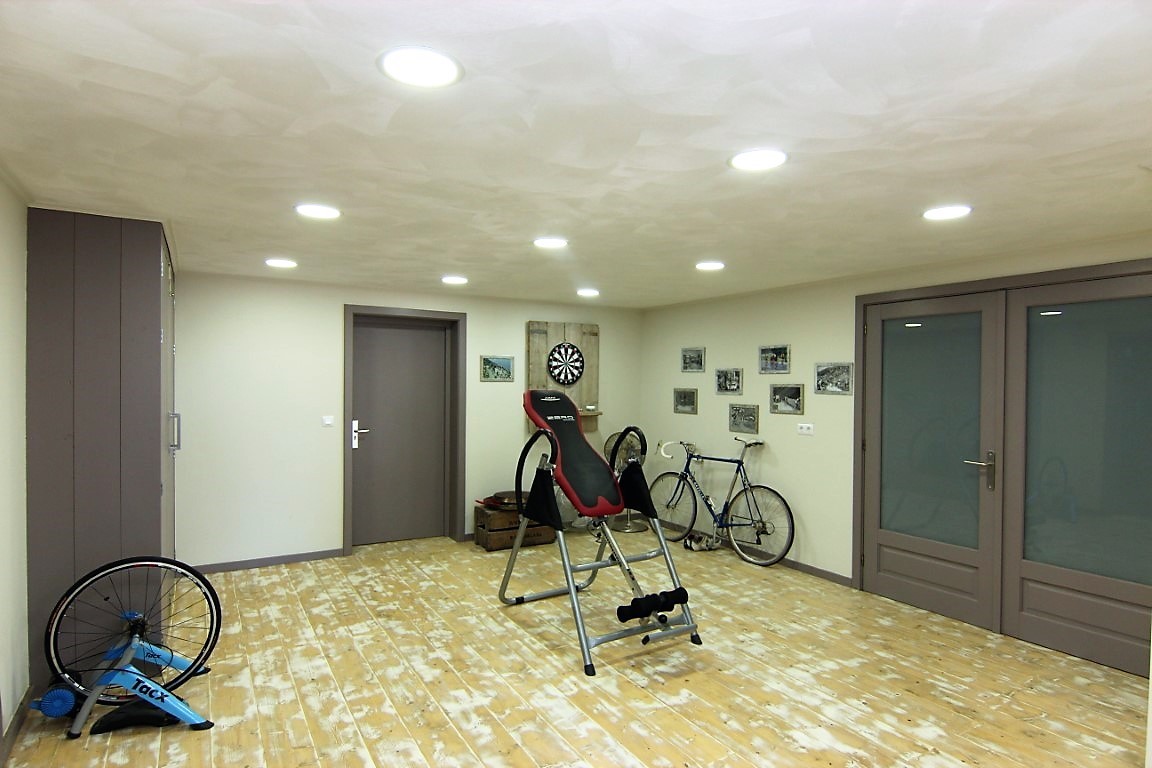 Fitnessraum im Untergeschoss mit hellem Holzboden, Deckeneinbauleuchten und Platz für Sportgeräte wie Fahrräder und eine Inversionsbank.