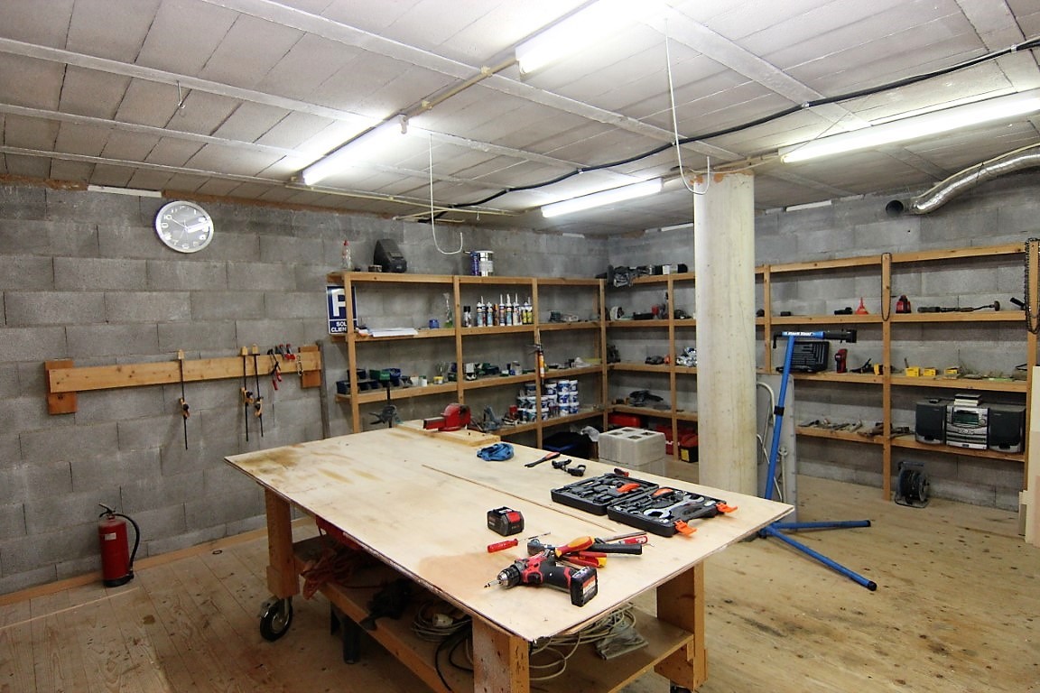 Geräumige Werkstatt im Keller mit Betonblockwänden, umlaufenden Holzregalen und einer großen zentralen Werkbank auf Holzboden.