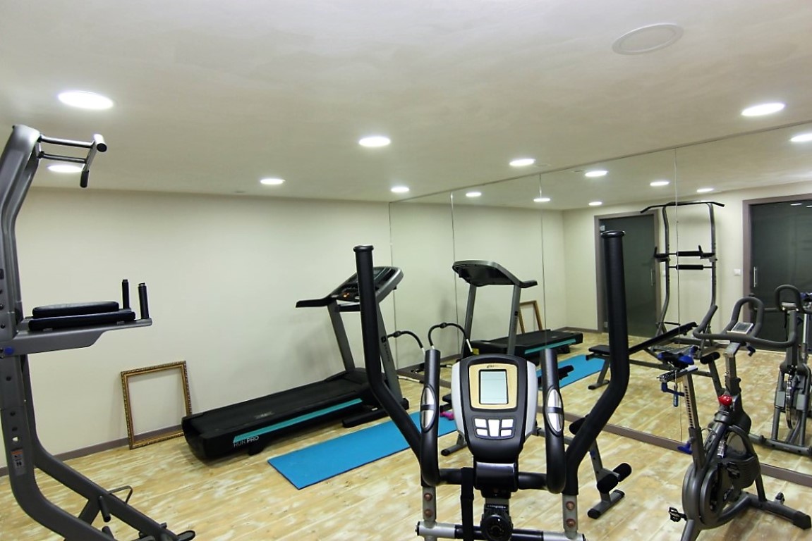 Privater Fitnessraum im Untergeschoss mit hellem Holzboden, wandhohen Spiegeln und diversen Trainingsgeräten unter modernen Deckeneinbaustrahlern.