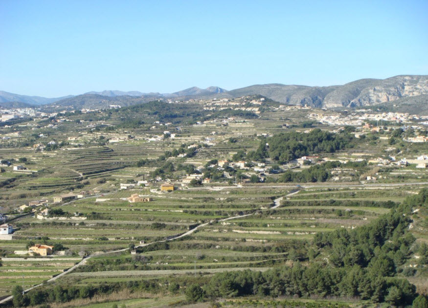 Panoramablick auf terrassiertes Gelände in Benitachell mit abgestufter Topographie, mediterraner Vegetation und Bergen im Hintergrund.