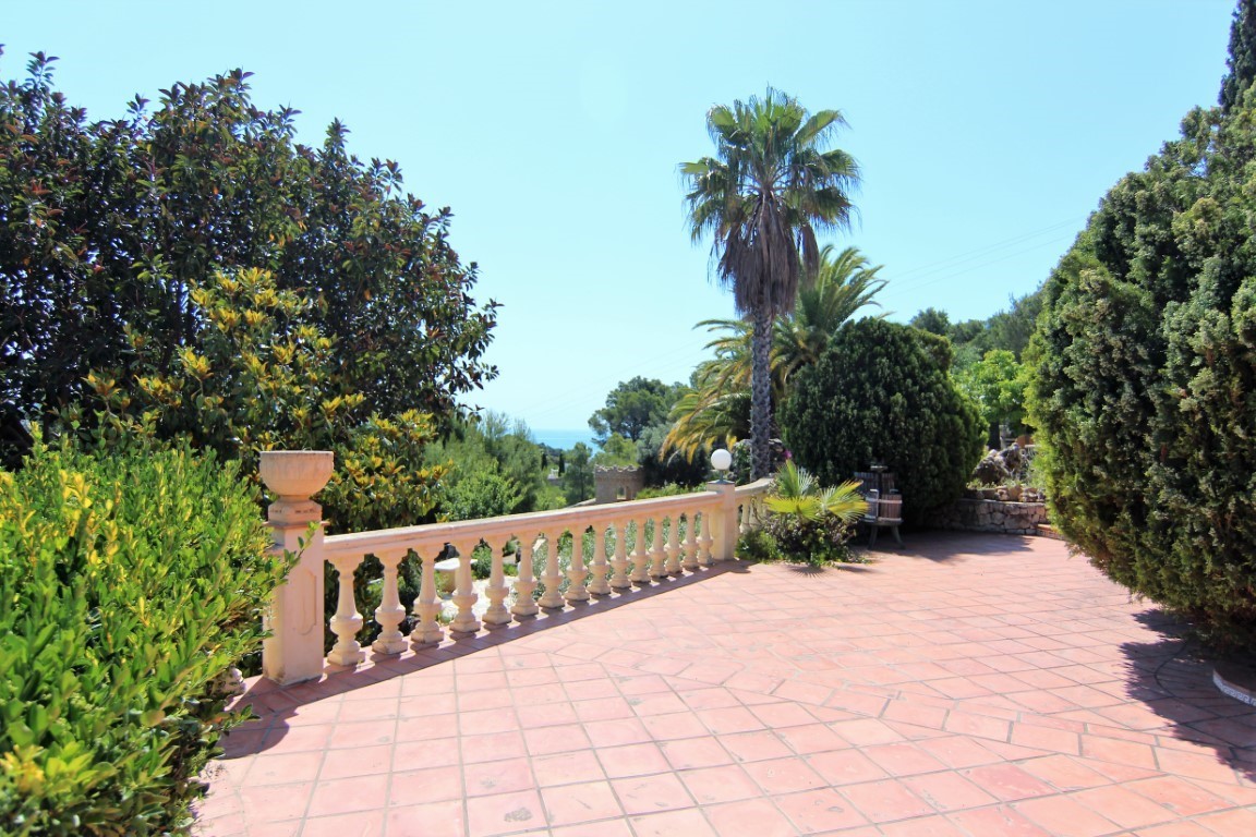 Außen-Terrasse mit Terrakotta-Fliesen und klassischer Steinbalustrade, umgeben von mediterraner Bepflanzung mit Blick auf das Meer in der Ferne.