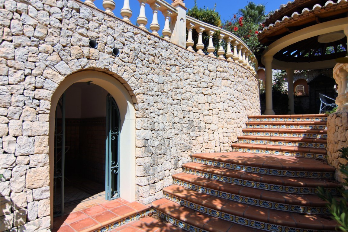 Außentreppe aus Terrakotta mit dekorativen Fliesen an einer geschwungenen Natursteinmauer. Mit Rundbogenzugang und klassischer Balustrade.