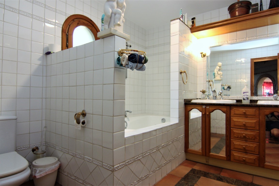 Badezimmer mit weiß gefliesten Wänden, Einbaubadewanne und Doppelwaschtisch aus Holz mit Granitplatte. Terrakottaboden und ein Rundbogenfenster ergänzen das Design.