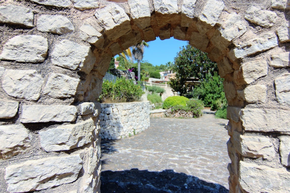 Ein Natursteinbogen rahmt den Eingang zu den mediterranen Gärten ein, mit gepflasterten Wegen und Trockensteinmauern unter blauem Himmel.