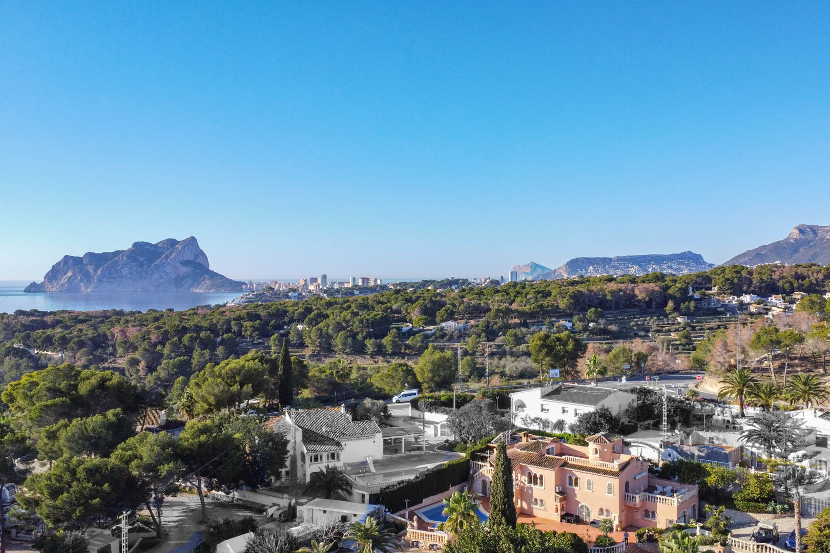 Erhöhter Panoramablick auf die Mittelmeerküste mit dem Peñón de Ifach, umgeben von dichten Pinienwäldern und Wohnvillen unter strahlend blauem Himmel.