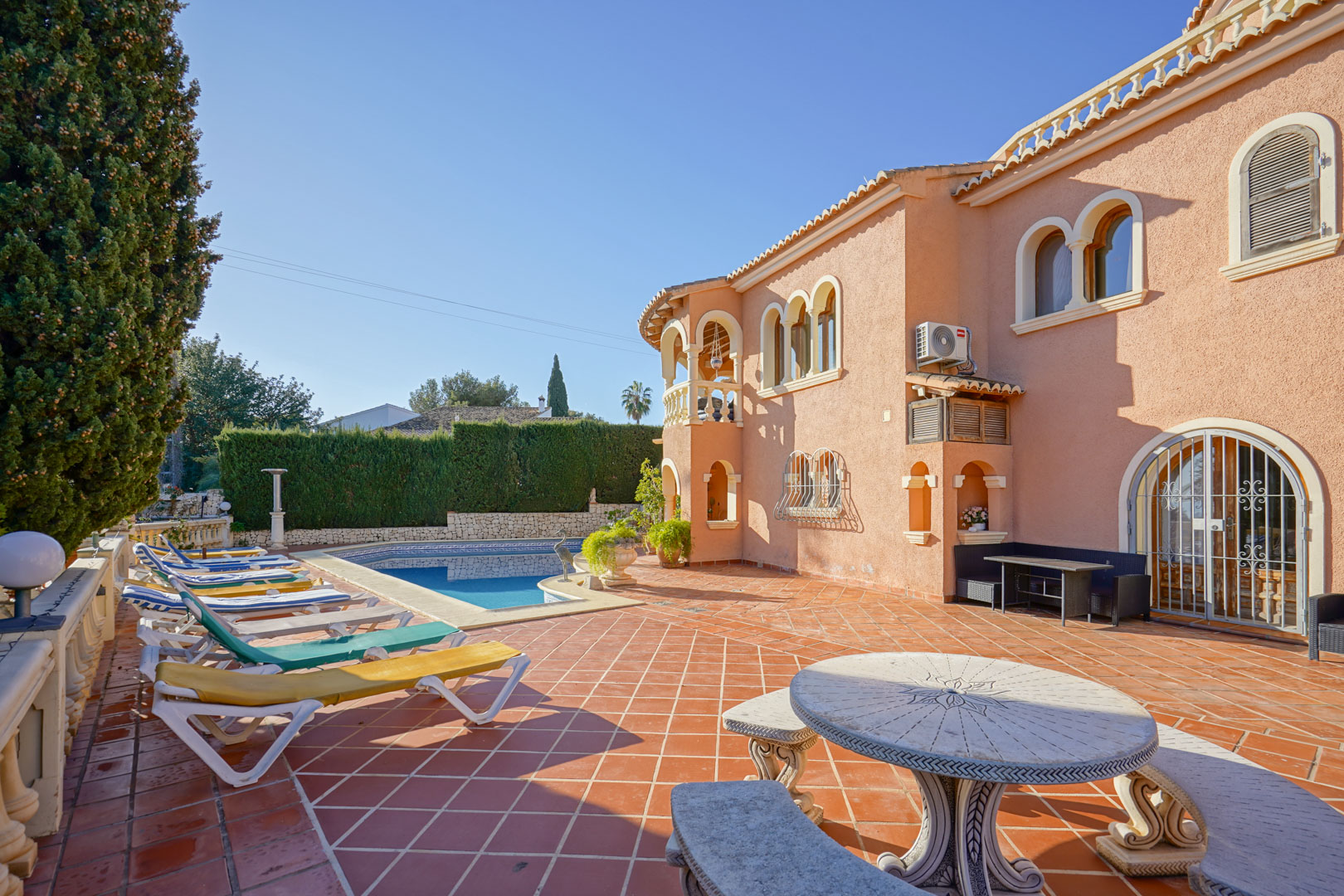 Großzügige Terrasse mit Terrakottafliesen, privatem Pool, Sonnenliegen und einem verzierten Steintisch vor einer mediterranen Fassade mit Rundbogenfenstern.