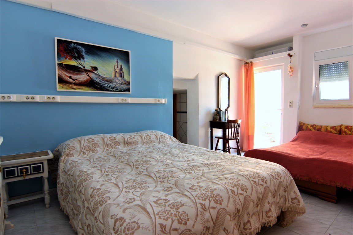 Helles Schlafzimmer mit blauer Akzentwand, Doppelbett mit gemusterter Decke und zusätzlichem Einzelbett. Mit Holzschreibtisch, Fliesenboden und direktem Zugang ins Freie.