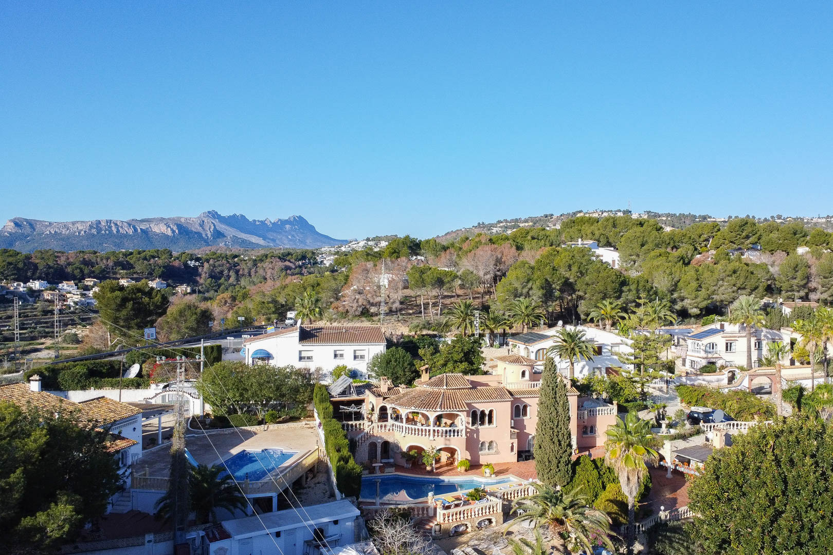 Luftaufnahme der mediterranen Villa mit geschwungenem Pool, Bogenterrassen und weitläufigem Garten vor einer markanten Bergkulisse.