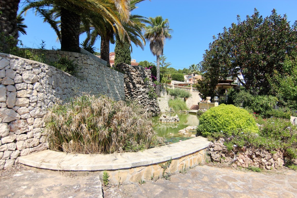 Mediterraner Garten mit Zierteich, Naturstein-Wasserfall und Trockensteinmauern, umgeben von Palmen und üppiger lokaler Vegetation.