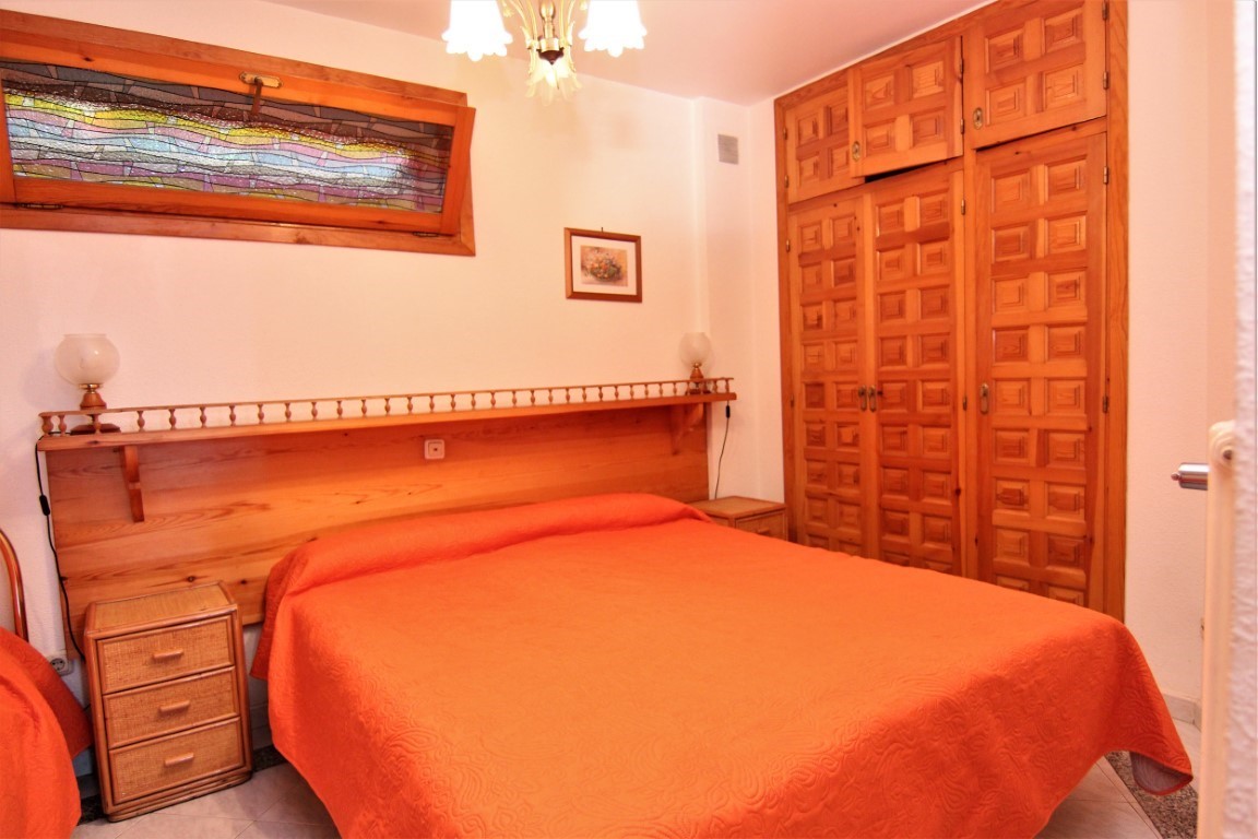 Schlafzimmer mit Doppelbett, integriertem Holzkopfteil und Einbauschränken. Ein horizontales Buntglasfenster und Nachttische mit Korbgeflecht setzen besondere gestalterische Akzente.