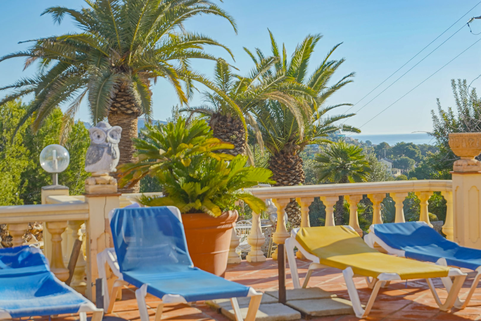 Sonnenterrasse mit Terrakottafliesen, klassischer Balustrade und Sonnenliegen, umgeben von hohen Palmen und mit weitem Blick auf das Mittelmeer.