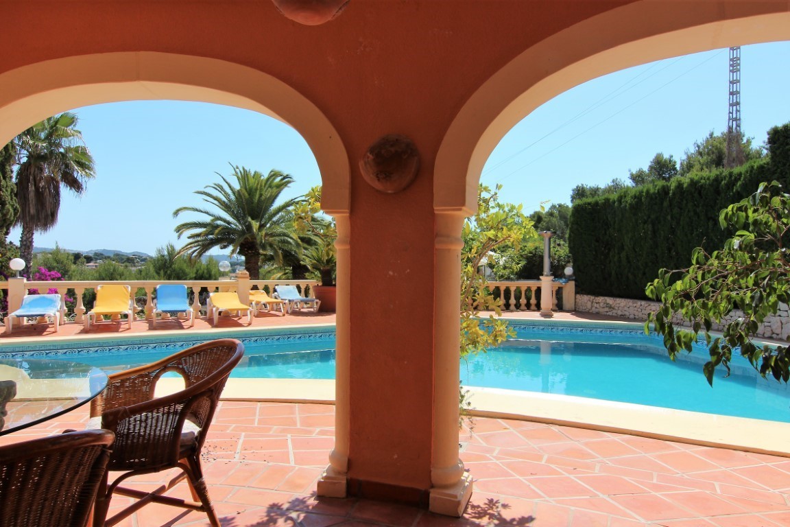 Überdachte Terrasse mit mediterranen Rundbögen und Blick auf den Poolbereich mit Sonnenliegen, umgeben von Palmen und gepflegten Hecken.