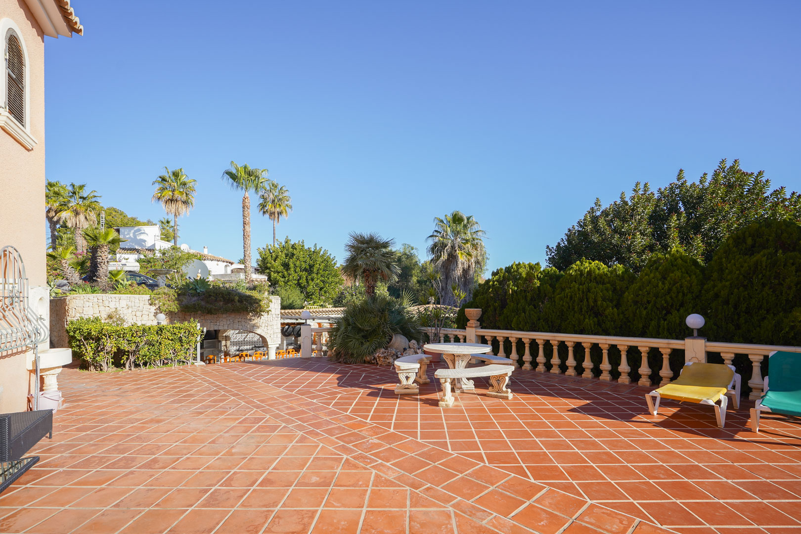 Weitläufige Außenterrasse mit Terrakottafliesen, Steinbalustrade und einer Sitzgruppe aus Stein, umgeben von Palmen und üppiger mediterraner Bepflanzung.