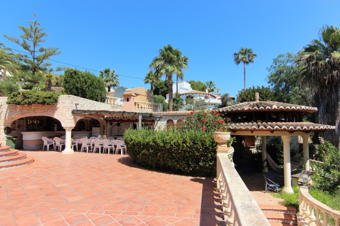 Weitläufige Terracotta-Terrasse mit einem überdachten Essbereich unter Steinbögen und einem rustikalen Pavillon, eingebettet in einen mediterranen Garten mit Palmen.