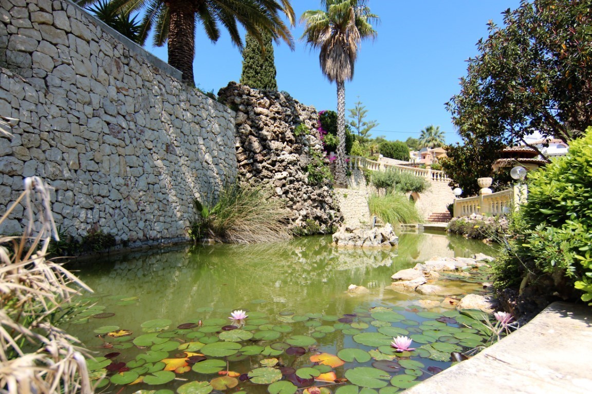 Zierteich mit Seerosen und Natursteinwasserfall, gesäumt von Trockenmauern und mediterraner Bepflanzung mit Palmen.