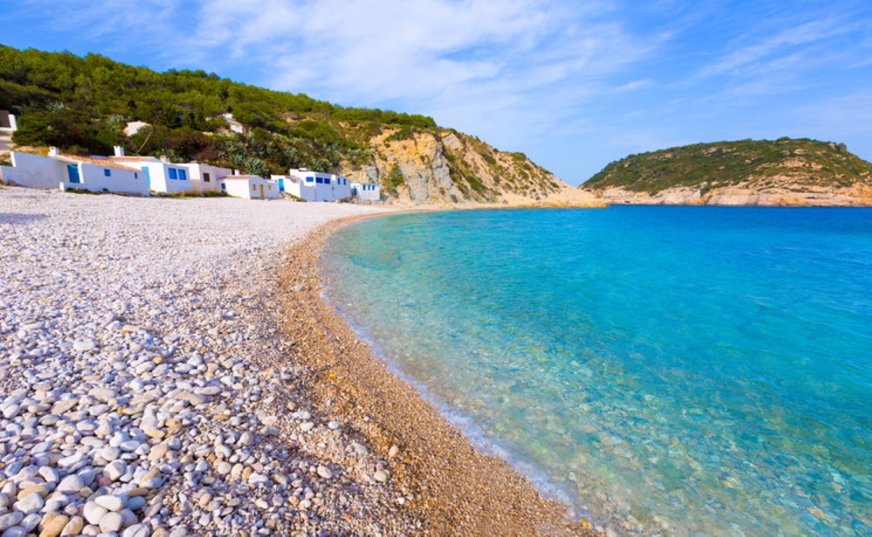 Blick auf die Cala de la Barraca mit türkisfarbenem Wasser, weißem Kieselstrand und traditionellen Fischerhäusern gegenüber der Insel Portichol in Jávea.