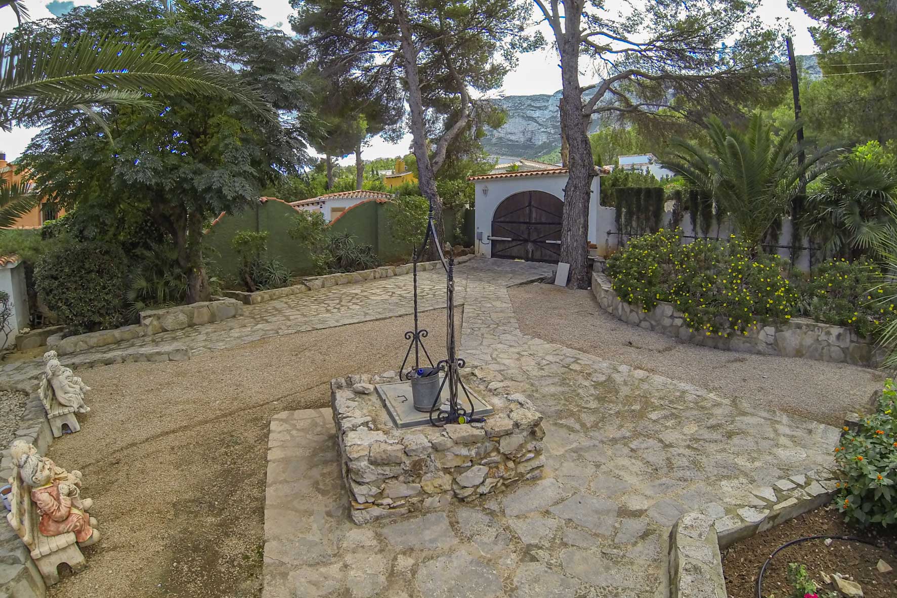 Grundstückseingang mit Steinbrunnen, gepflastertem Weg und Holztor im Hintergrund.