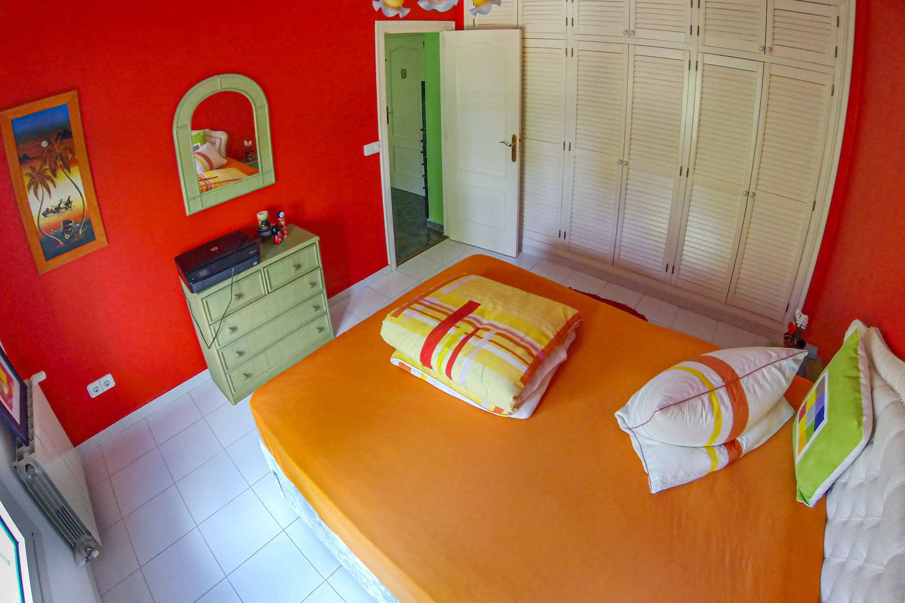 Schlafzimmer mit roten Wänden, weißem Einbauschrank und einem Bett mit orangefarbener Bettwäsche.
