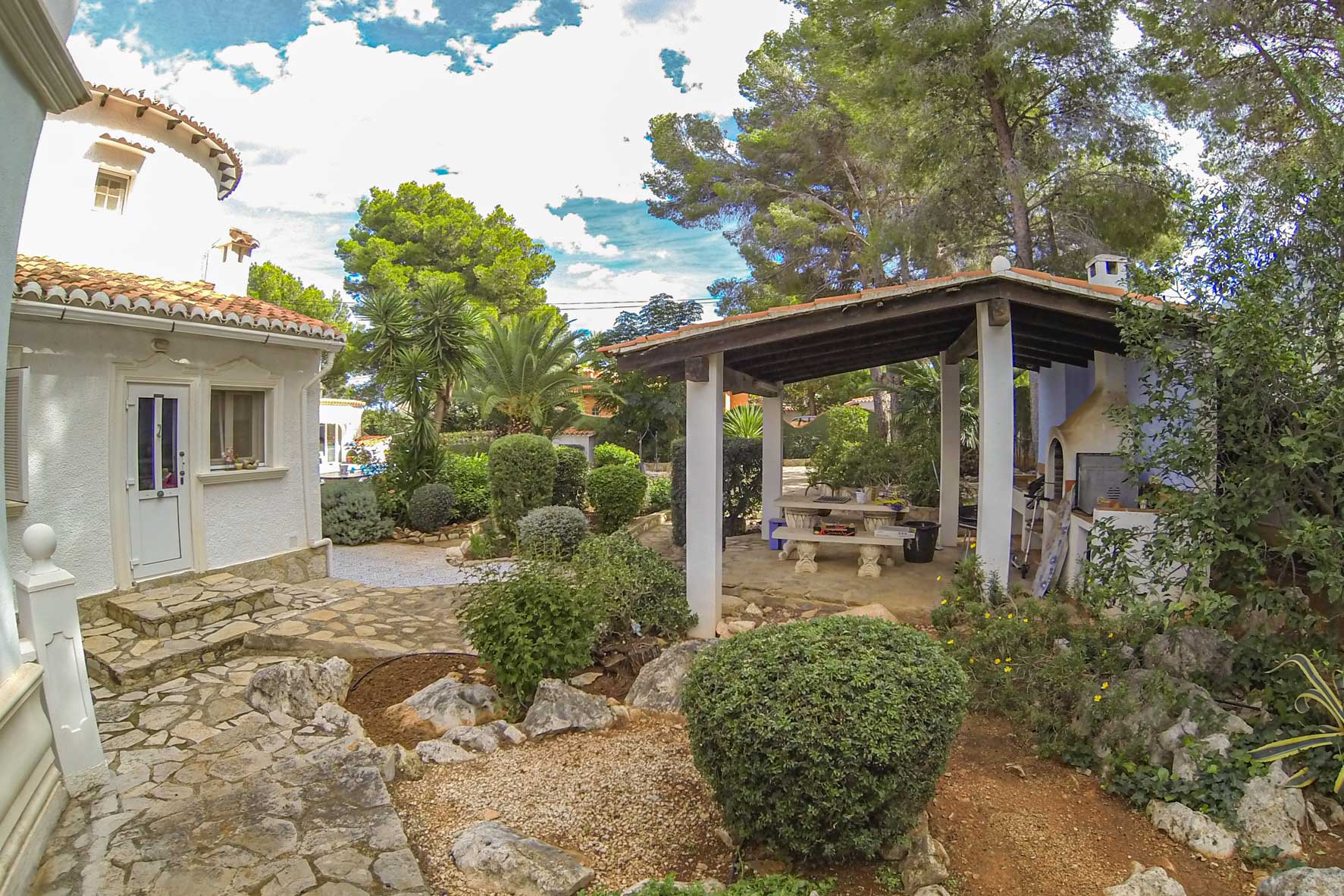 Überdachter Grillplatz mit Holzofen und Steintisch in einem mediterranen Garten.