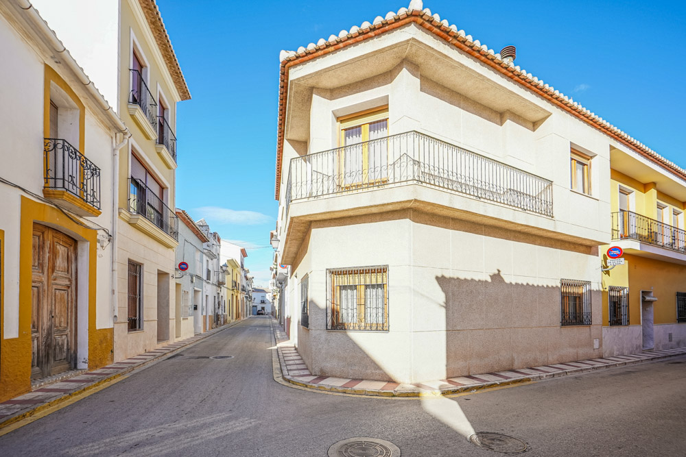 Außenansicht des Eck-Stadthauses mit weißer Fassade, umlaufendem Balkon mit Eisenbeschlägen und traditionellen Fenstern in einer ruhigen Gasse von Gata de Gorgos.