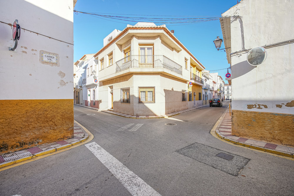 Außenansicht eines traditionellen Eckhauses in Gata de Gorgos mit umlaufendem Balkon, schmiedeeisernen Geländern und klassischen Fenstergittern in einer ruhigen Straße.