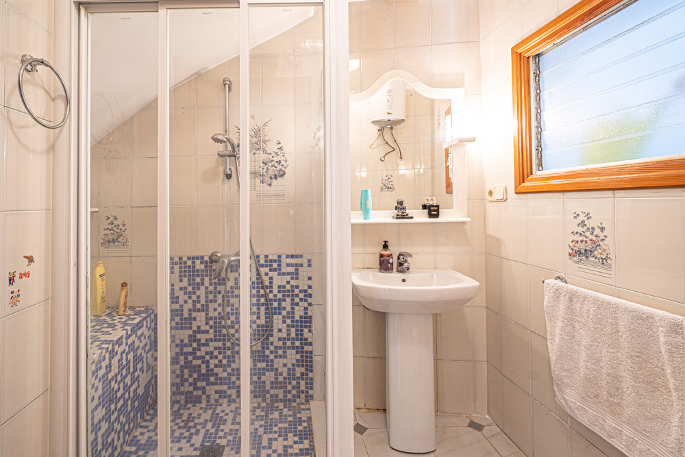 Badezimmer mit einer begehbaren Dusche aus blauem Mosaik, einem Säulenwaschbecken und gefliesten Wänden mit floralen Akzenten und Holzfensterrahmen.