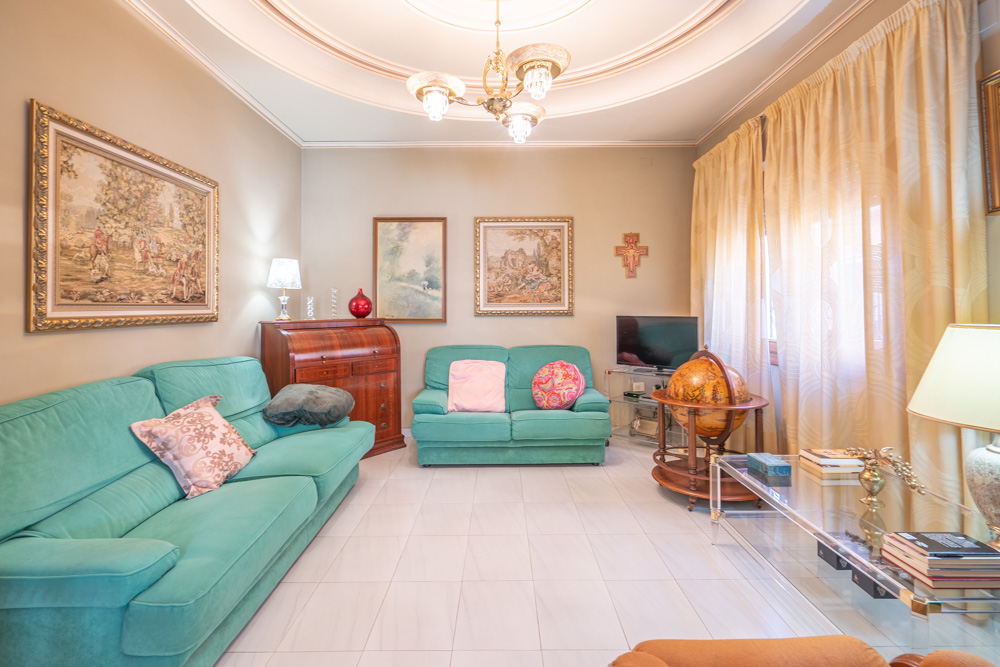 Helles Wohnzimmer mit türkisfarbenen Sofas, weißen Fliesenböden und kreisförmigem Deckenstuck. Ausgestattet mit klassischen Holzmöbeln, einem Sekretär und gerahmten Wandbildern.