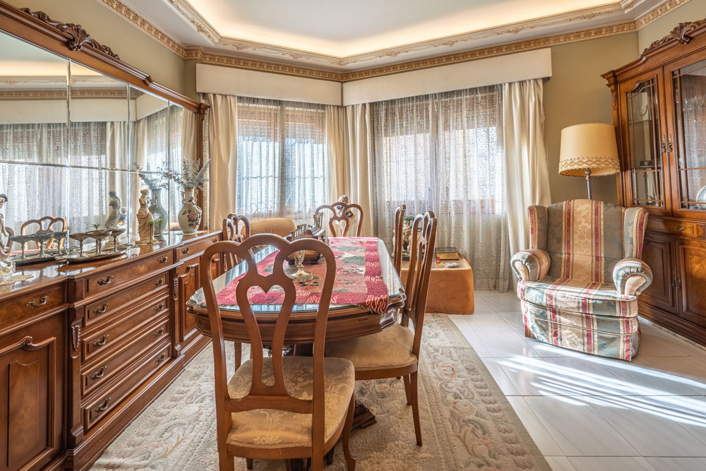Klassisches Esszimmer mit geschnitzten Holzmöbeln, Glastisch, verspiegeltem Sideboard und dekorativen Deckenleisten mit indirekter Beleuchtung.