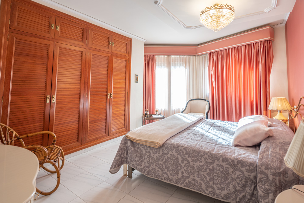 Schlafzimmer mit massiven Holzeinbauschränken, großem Fenster mit rosa Vorhängen, Fliesenboden und einem eleganten Kristallkronleuchter.