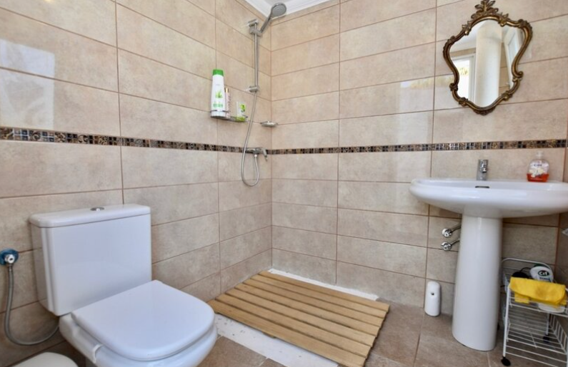 Badezimmer mit begehbarer Dusche, Holzlattenrost, beigen Fliesen mit Mosaikbordüre und einem verzierten Spiegel mit Goldrahmen.