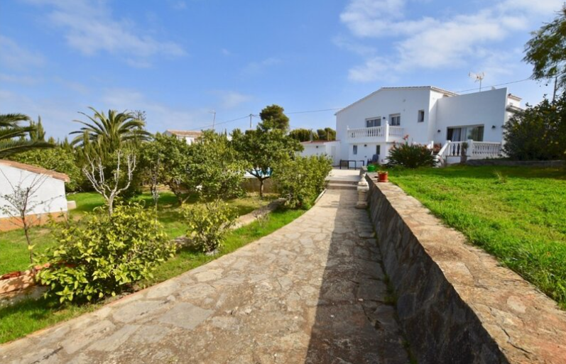 Ein Natursteinpfad führt durch den mediterranen Garten zur weißen Villa, gesäumt von gepflegten Rasenflächen, Palmen und traditionellen Steinmauern.