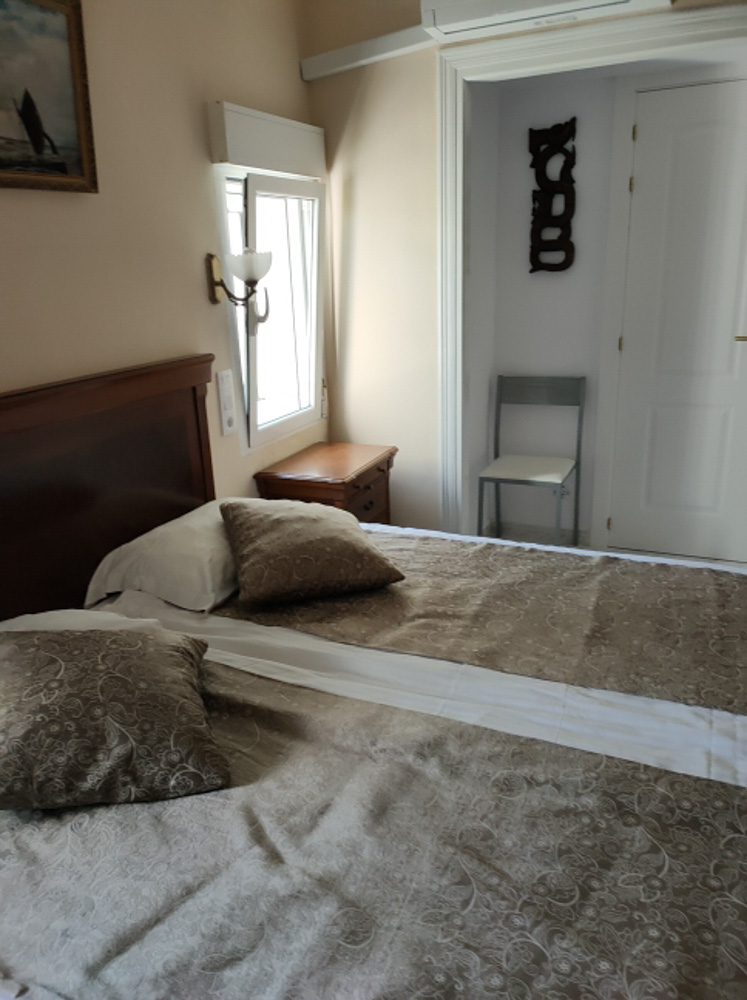 Schlafzimmer mit Doppelbett, klassischem Holzkopfteil und Klimaanlage. Es bietet natürliches Licht durch ein Seitenfenster, einen Nachttisch und einen praktischen Sitzbereich.