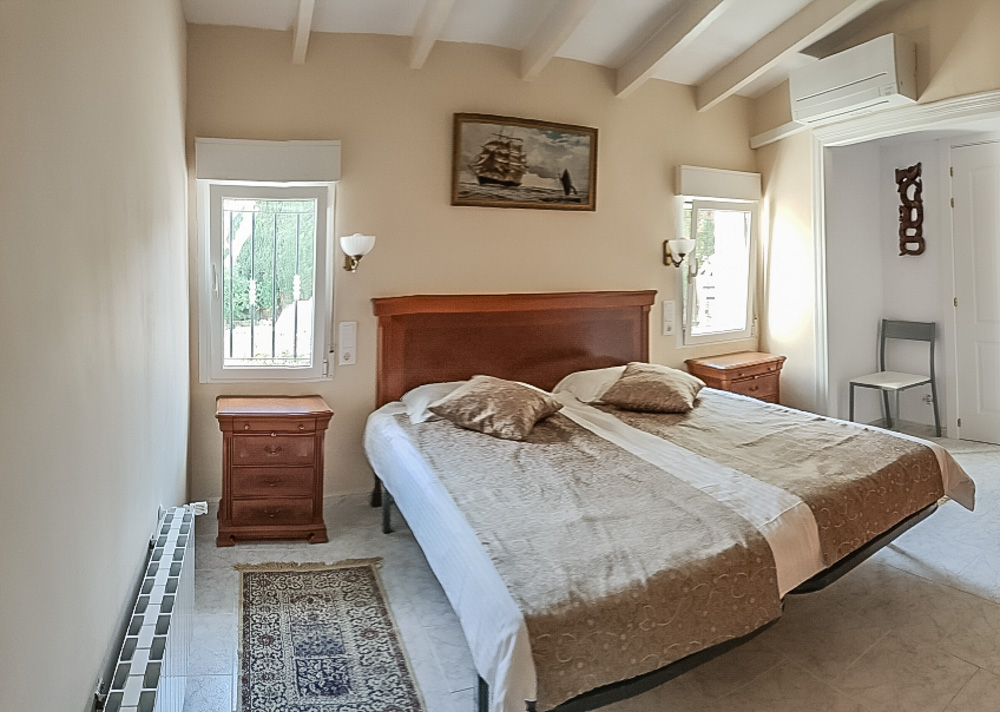 Schlafzimmer mit weißer Balkendecke, Doppelbett mit Holz-Kopfteil, passenden Nachttischen, Klimaanlage und Heizkörper.