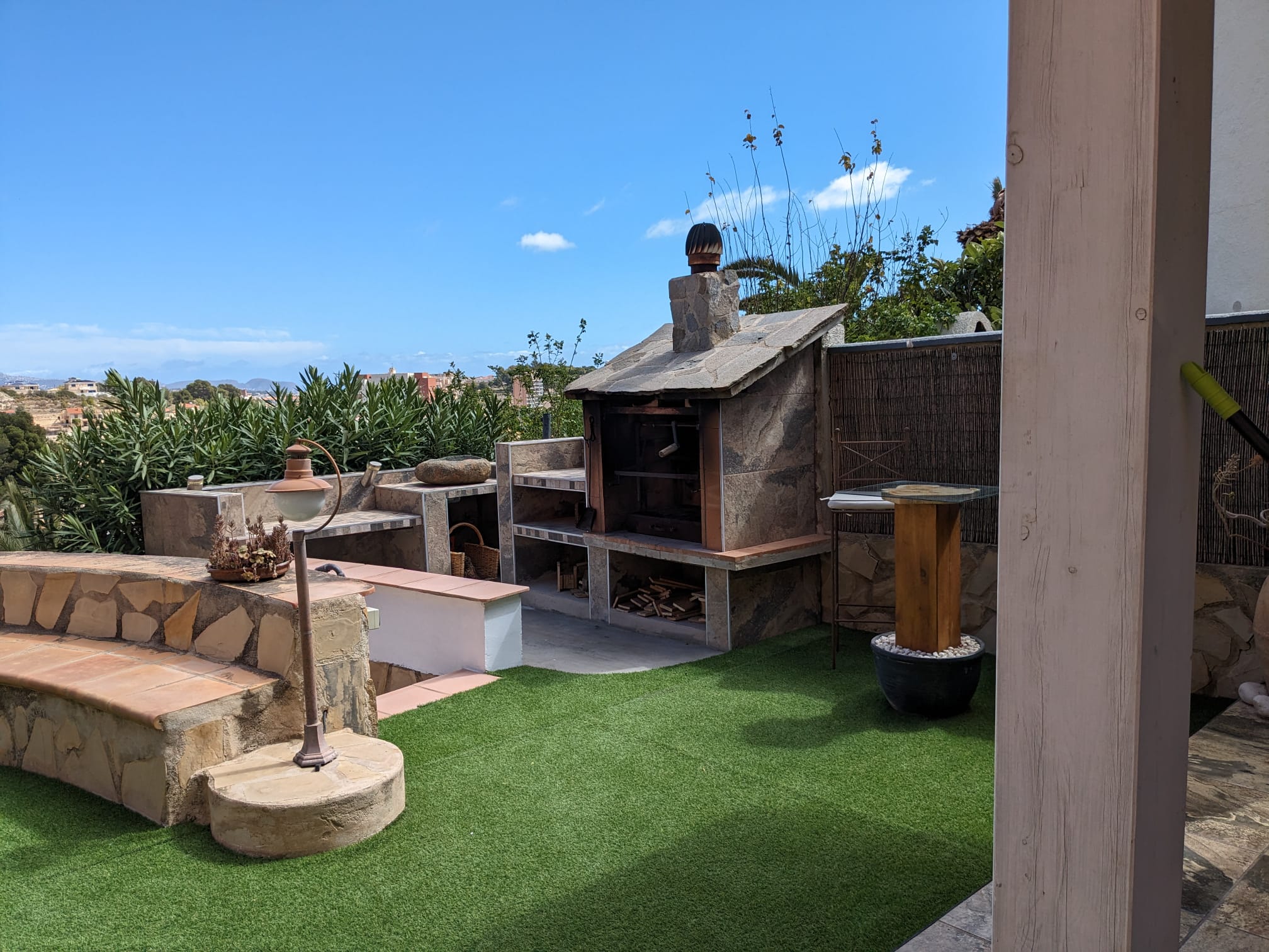 Außenbereich mit gemauertem Naturstein-Grill, integrierten Arbeitsflächen, einer Steinbank und Kunstrasen unter strahlend blauem Himmel.