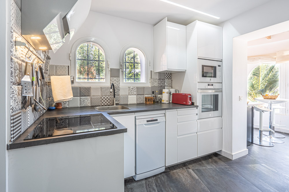 Moderne Küche mit weißen Hochglanzschränken, dekorativem Fliesenspiegel und integrierten Geräten. Rundbogenfenster bieten natürliches Licht und Blick auf die Gartenanlage.