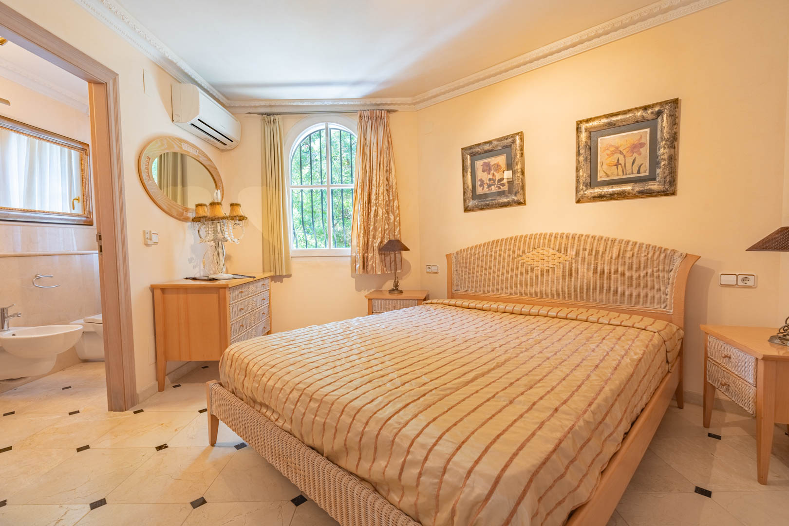 Schlafzimmer mit Korbbett, Marmorboden mit schwarzen Akzenten und angrenzendem Badezimmer. Ausgestattet mit Klimaanlage, Zierleisten und einem Rundbogenfenster.