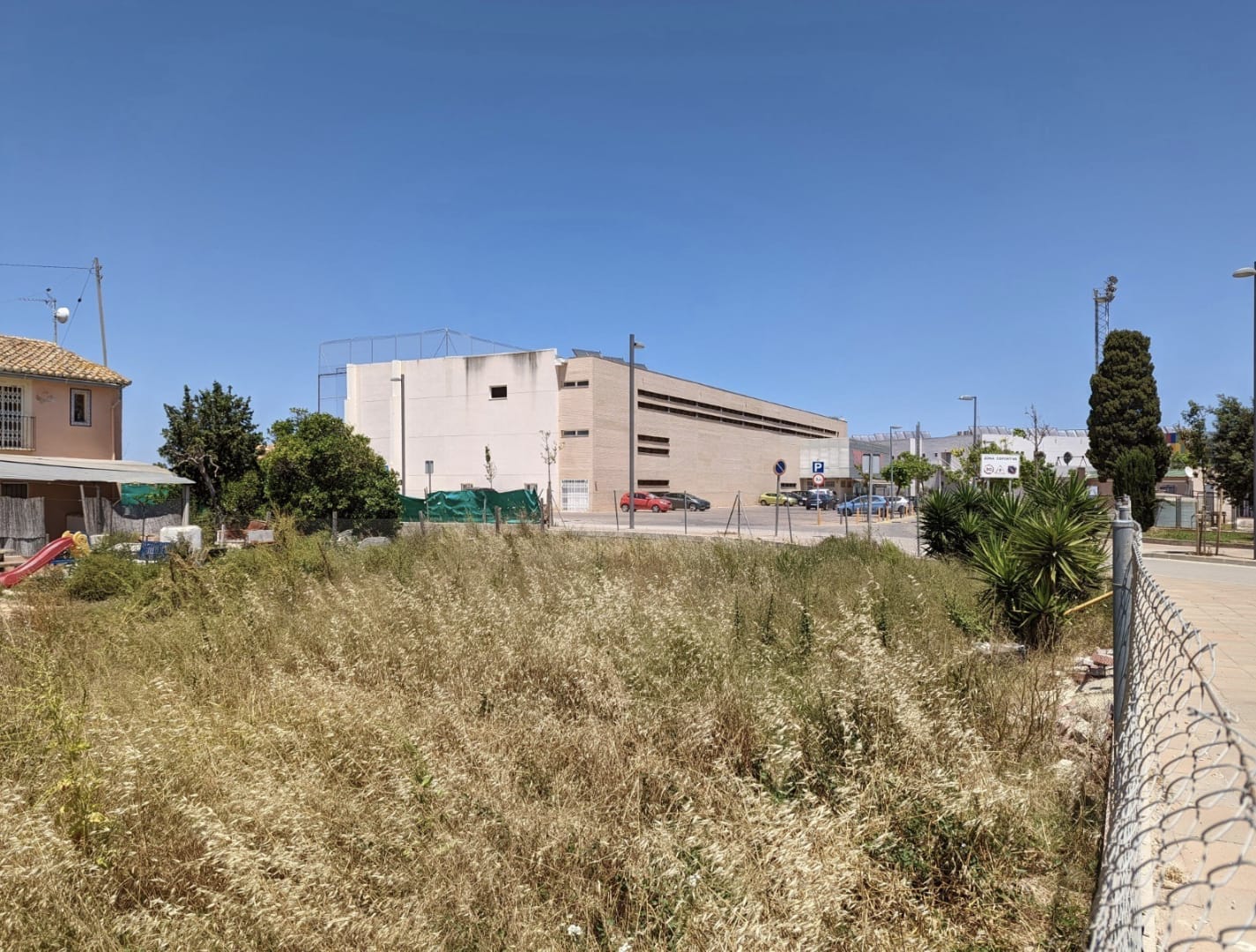 Ebenes Stadtgrundstück mit natürlicher Vegetation, direkt gegenüber einer modernen Sportanlage und öffentlichen Parkplätzen gelegen.