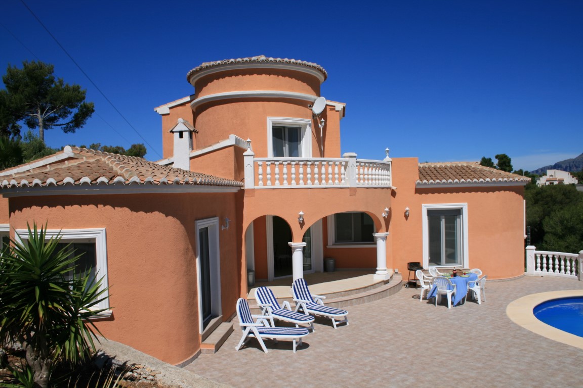 Außenansicht einer mediterranen Villa mit terracottafarbener Fassade, Rundturm-Elementen, großzügiger Fliesenterrasse, Sonnenliegen und privatem Poolbereich.
