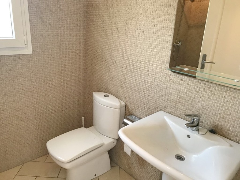 Badezimmer mit beigefarbenen Mosaikfliesen, weißem Keramikwaschbecken und passendem WC. Ein Spiegel mit Glasablage und neutrale Bodenfliesen prägen den Raum.