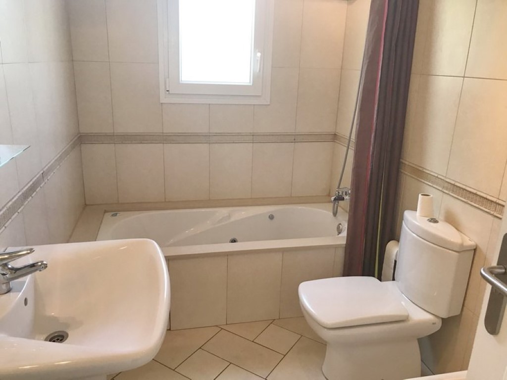 Badezimmer mit eingebauter Whirlpool-Badewanne, weißem Keramikwaschbecken und WC. Beige Wandfliesen mit Zierborte und helles, diagonal verlegtes Bodenmuster.