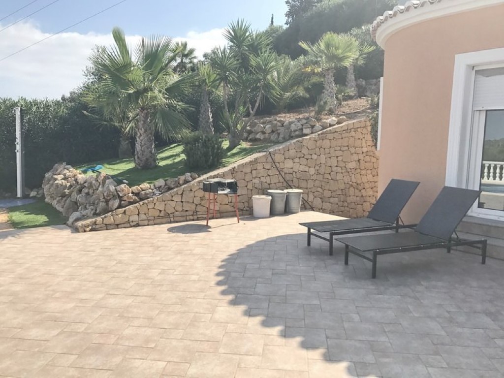 Gepflasterte Terrasse mit Sonnenliegen und Grill, begrenzt durch eine Natursteinmauer und einen mediterranen Garten mit Palmen auf einer Anhöhe.