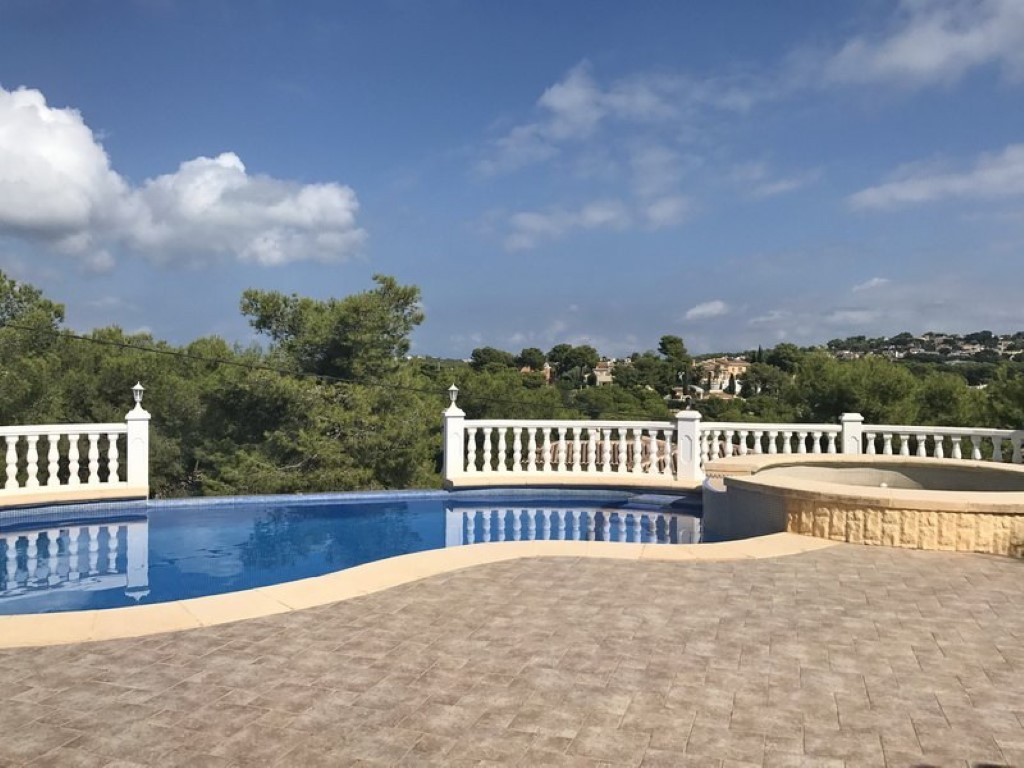 Geschwungener Pool mit angrenzendem Whirlpool, umgeben von einer gepflasterten Terrasse und weißer Balustrade mit Blick auf die mediterrane Landschaft.
