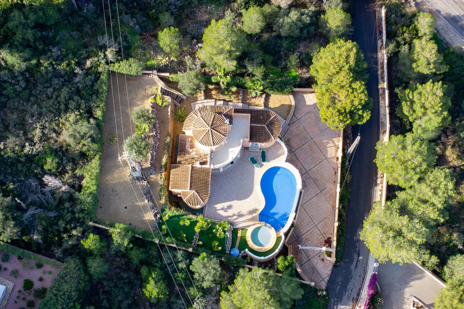 Luftaufnahme der Villa mit Terrakotta-Ziegeldächern, nierenförmigem Pool und kreisförmigem Whirlpool, umgeben von einem dichten mediterranen Waldgebiet.