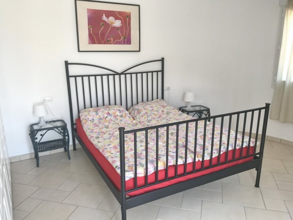 Schlafzimmer mit schwarzem Schmiedeeisenbett, passenden Nachttischen aus Korbgeflecht und hellem Fliesenboden. Natürliches Licht erhellt die weißen Wände.