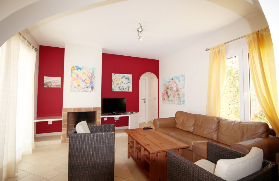 Wohnzimmer mit roter Akzentwand, traditionellem Backsteinkamin, braunem Ledersofa und dunklen Korbsesseln auf hellem Fliesenboden mit natürlichem Lichteinfall.