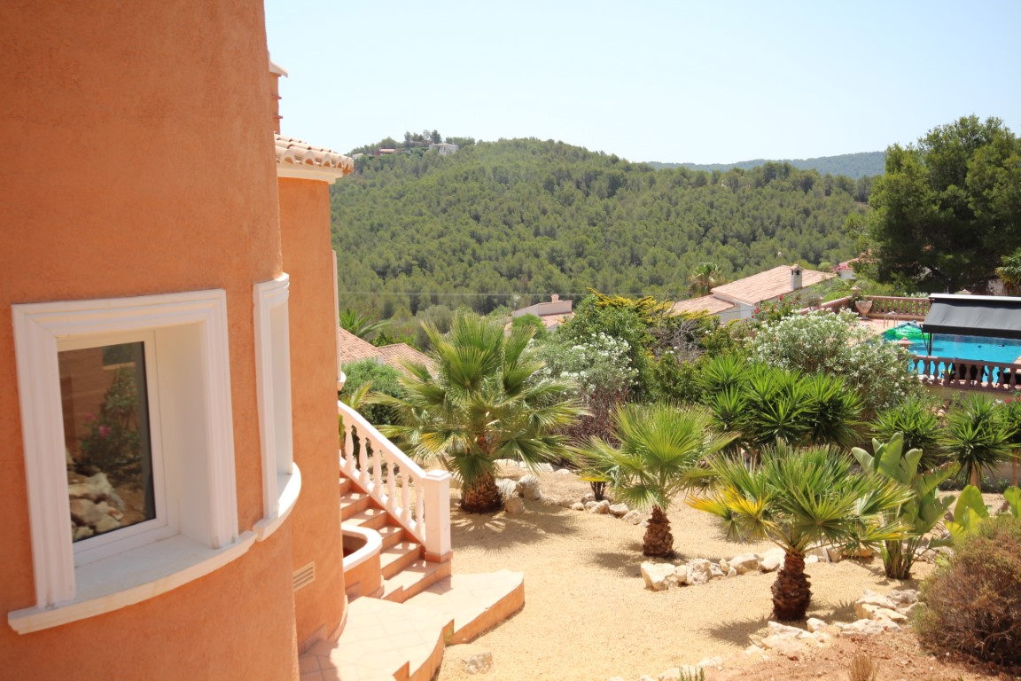 Außenansicht der terrakottafarbenen, geschwungenen Fassade mit pflegeleichtem mediterranem Garten, Palmen und weitem Blick über das bewaldete Tal.