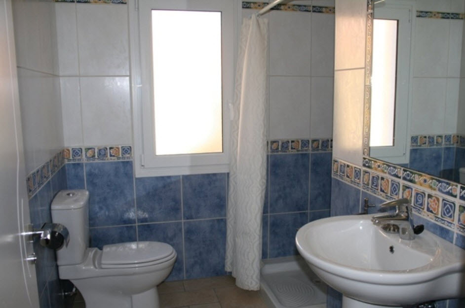 Badezimmer mit blau-weißen Fliesen, dekorativer Bordüre, Säulenwaschbecken, Duschwanne mit Vorhang und einem Milchglasfenster für natürliches Licht.