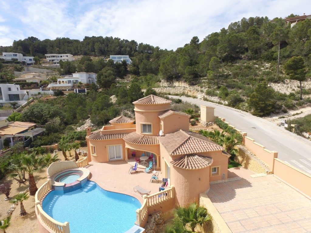 Luftaufnahme der mediterranen Villa mit terracottafarbener Fassade, geschwungenem Pool mit integriertem Jacuzzi und weitläufiger Sonnenterrasse inmitten grüner Pinienwälder.