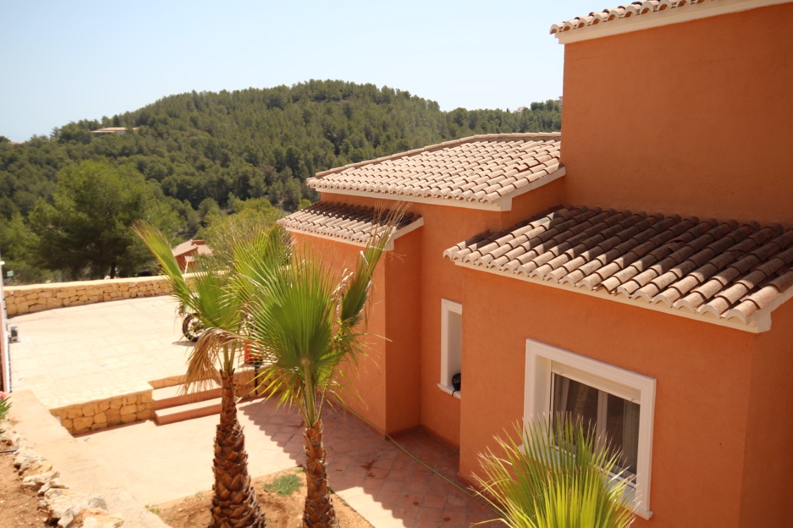 Mediterrane Fassade in Terrakotta-Tönen mit traditionellen Ziegeldächern, einem gepflasterten Innenhof mit Palmen und Blick auf die bewaldeten Hügel.